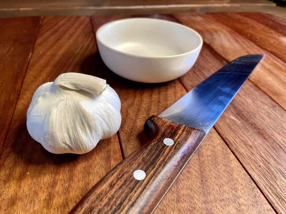 Head of garlic, knife, and bowl on chopping board.