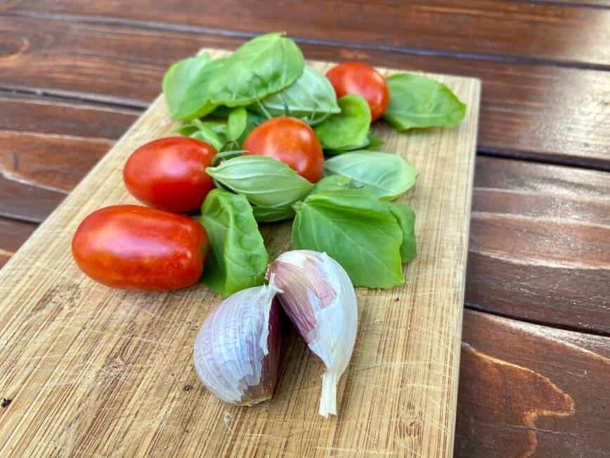 Tomatoes, fresh basil, and garlic cloves on a small wooden cutting board.