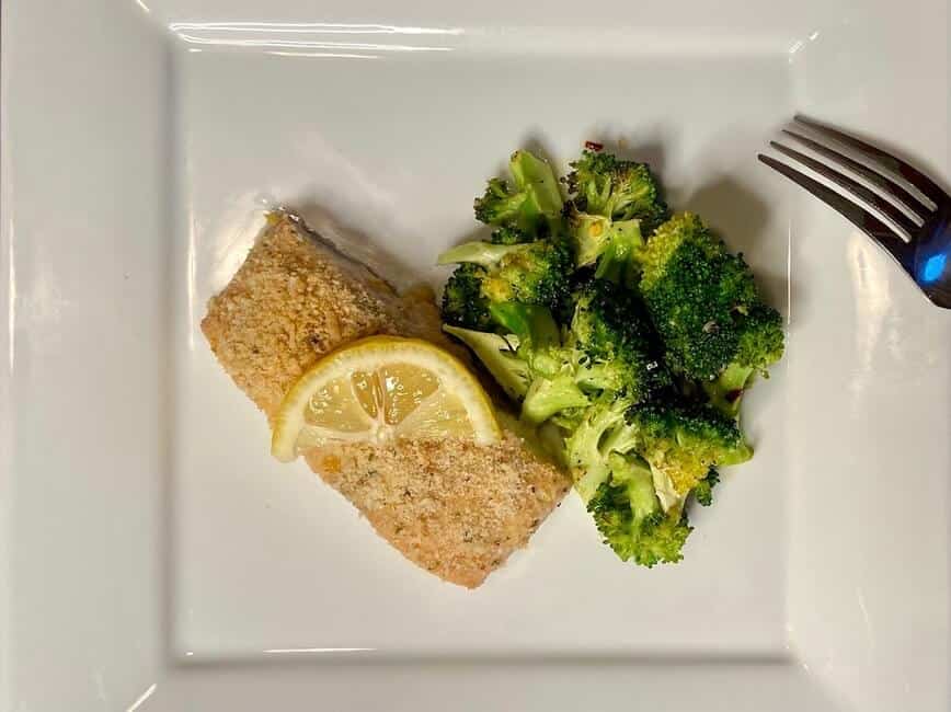 Salmon with a lemon wedge on top, next to broccoli on a plate.