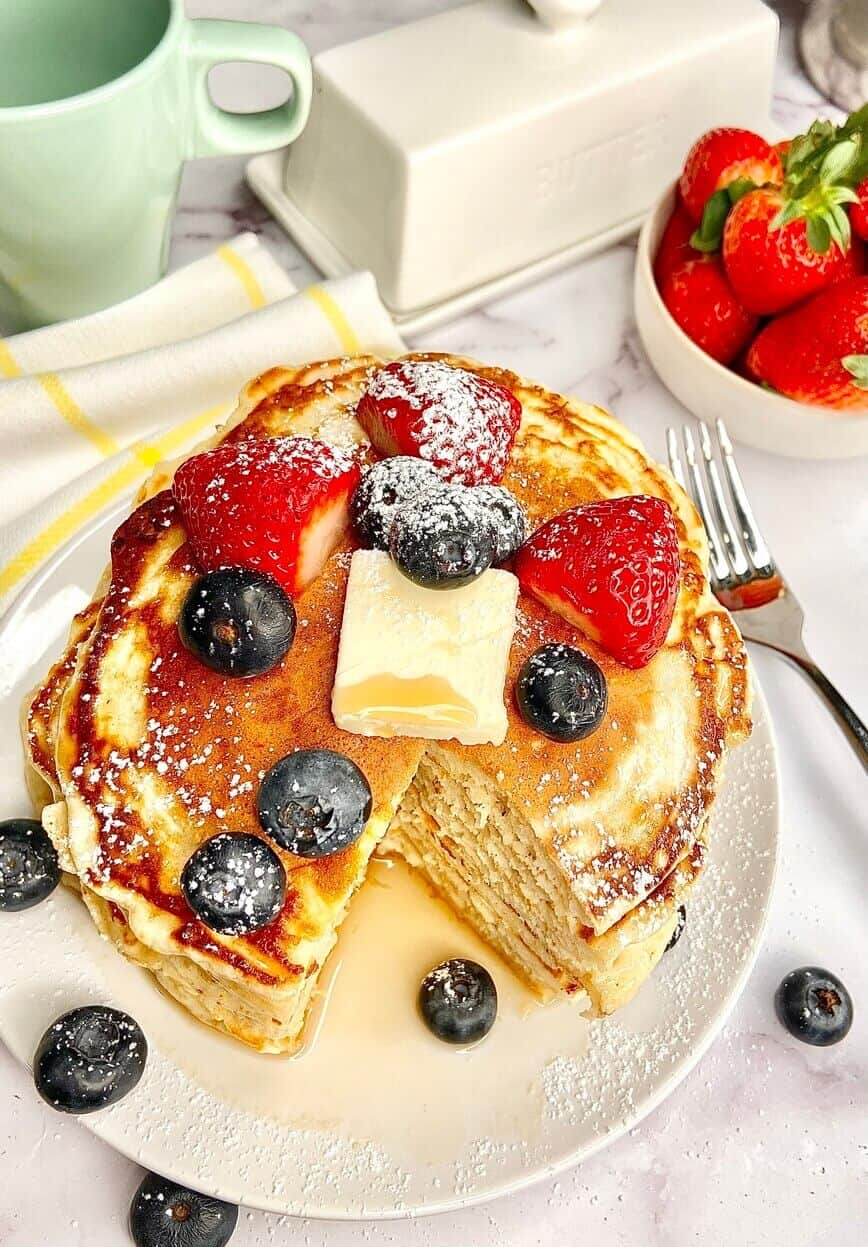 Stack of pancakes on a plate, with a bowl of berries, a dish of butter, and a cup of coffee.
