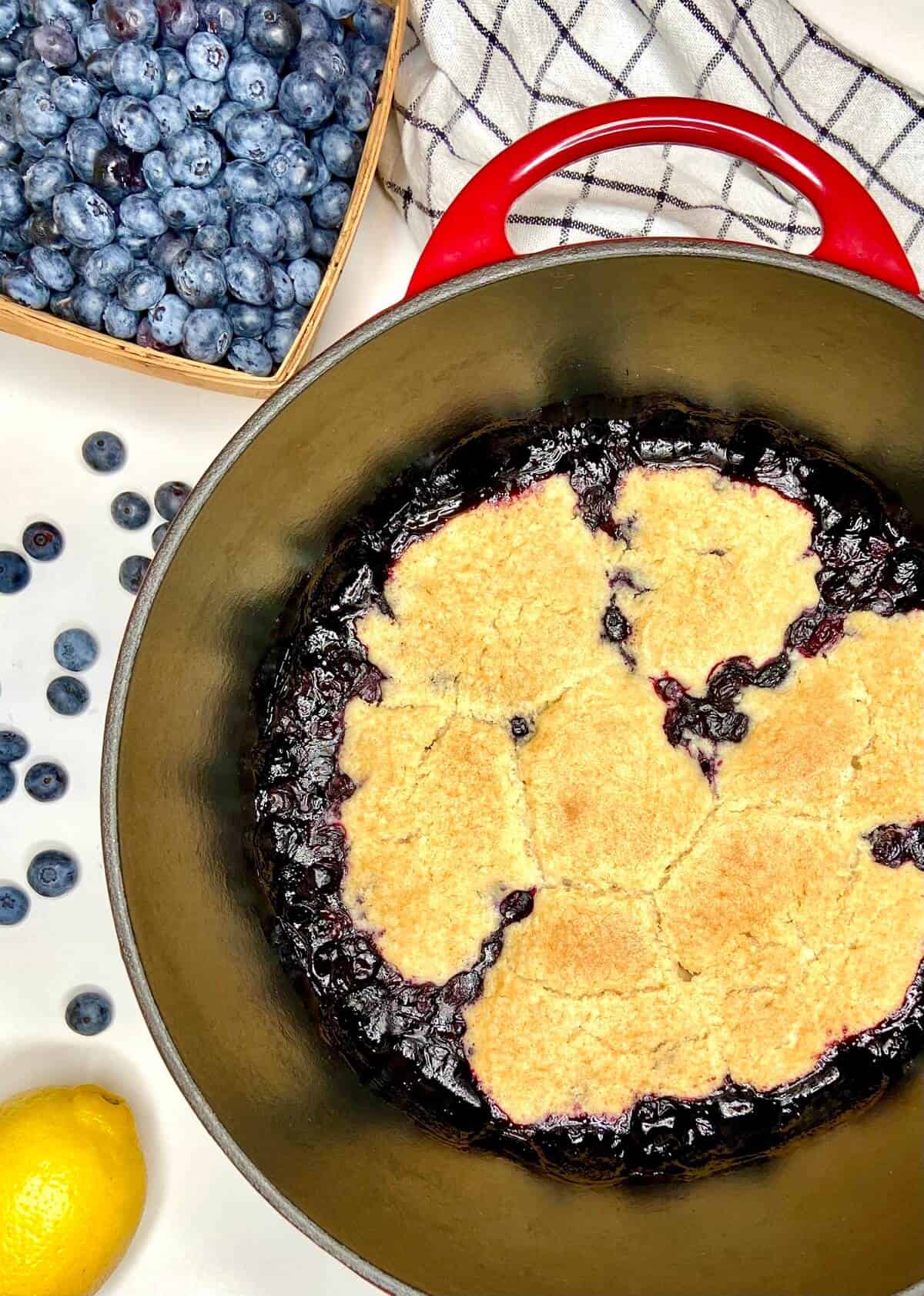 Cobbler in dutch oven, with a pint of blueberries and a towel on a white table.