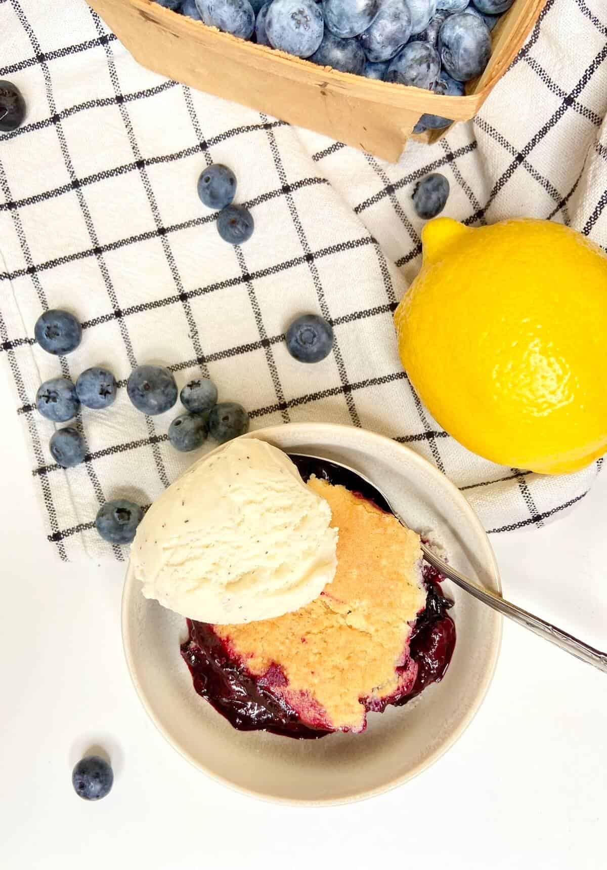 Cobbler with ice cream in a small dessert bowl, on a table with blueberries and a lemon.