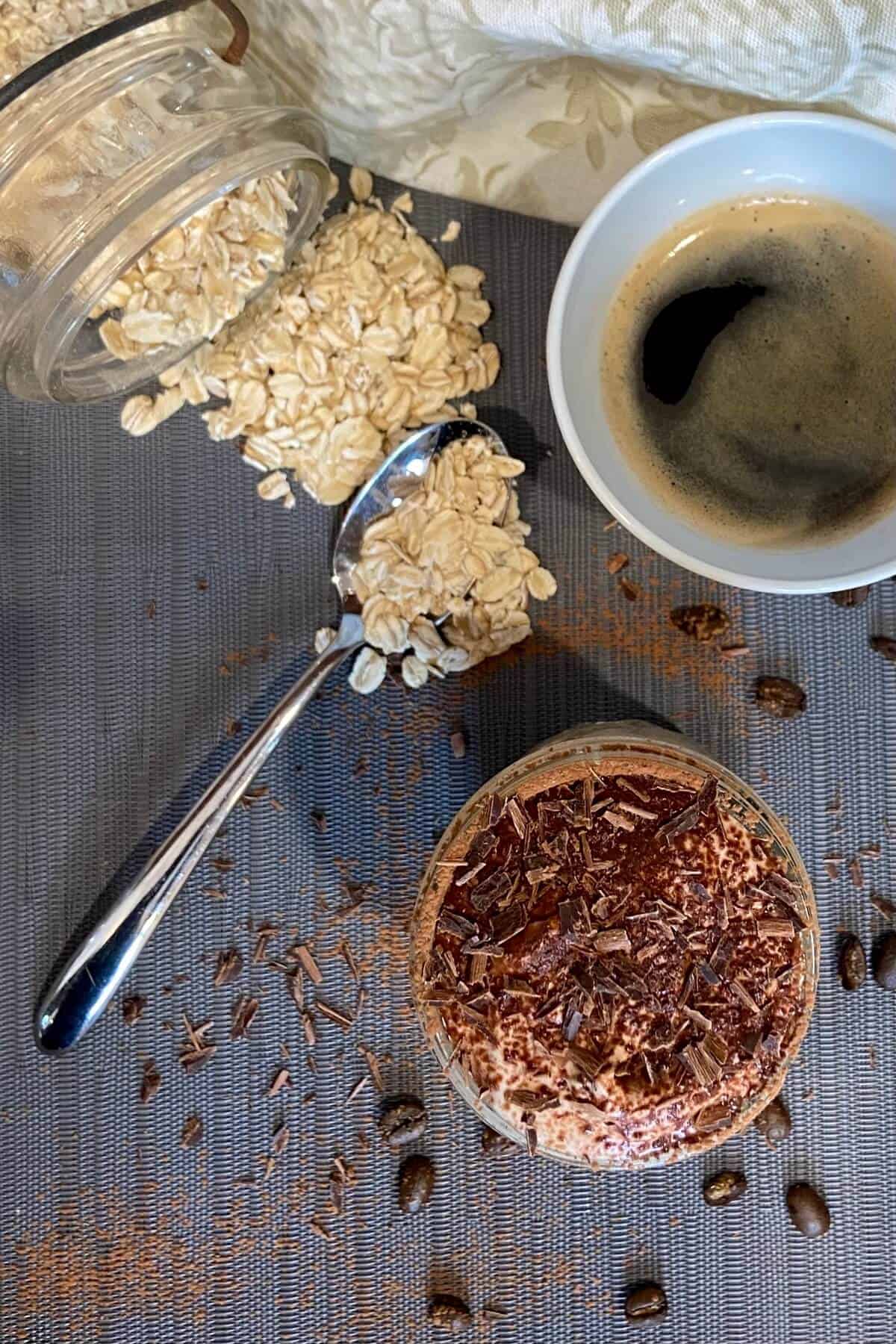 A jar of oats spilled onto a table with a cup of coffee and the overnight oats.