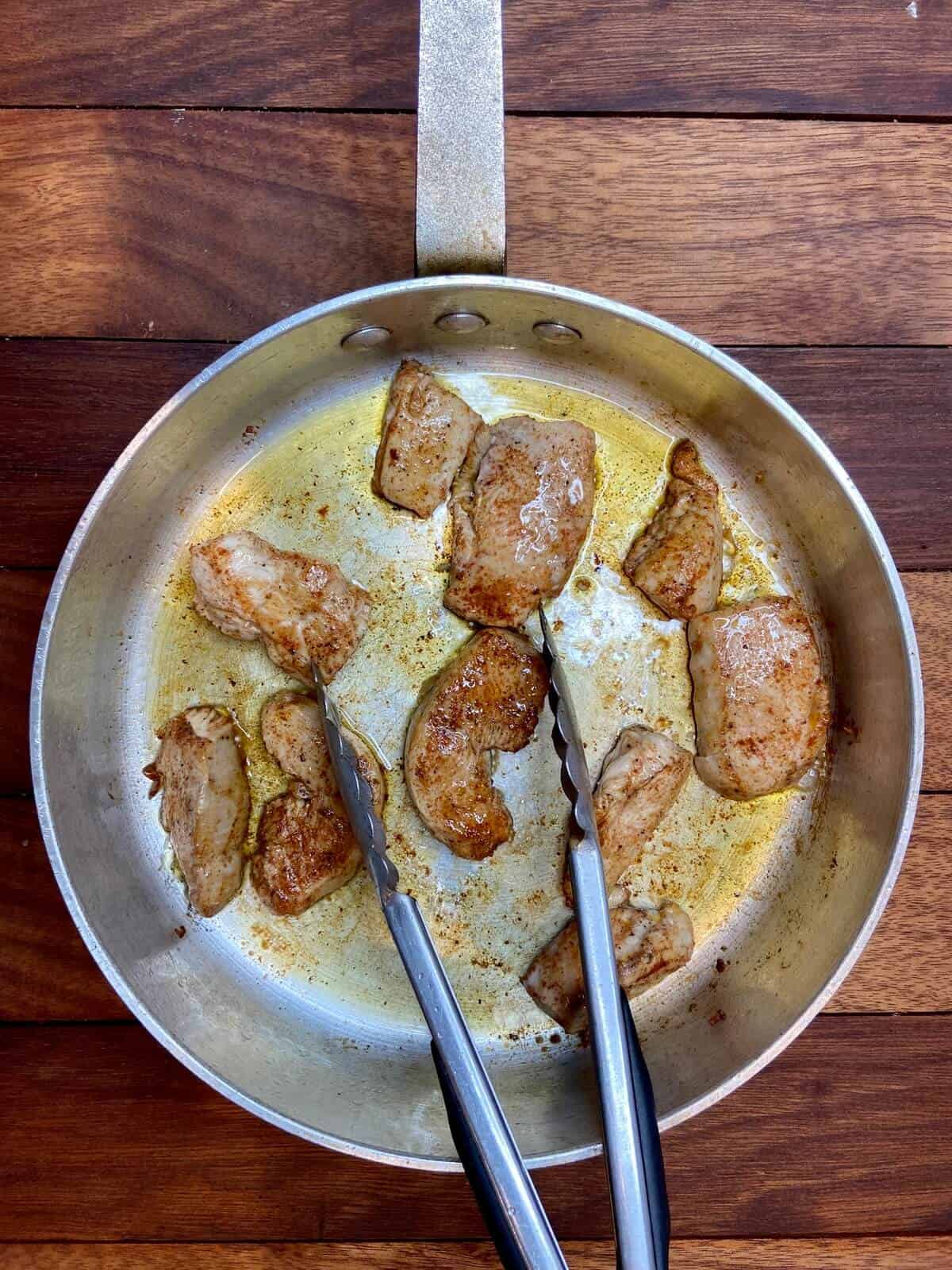 Chicken cooking in pan, with tongs.