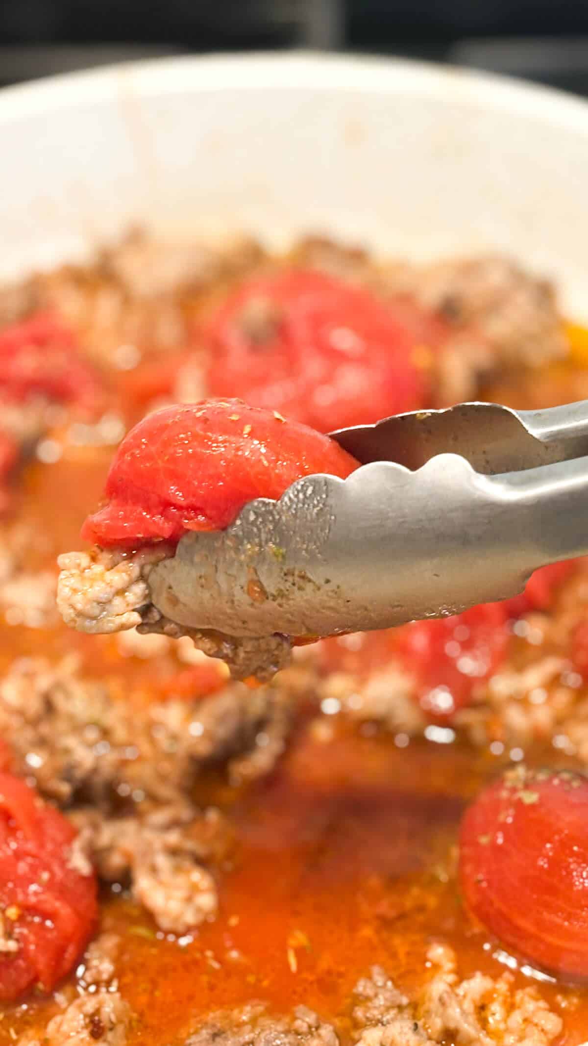 Breaking up plum tomatoes with tongs while simmering in pan.